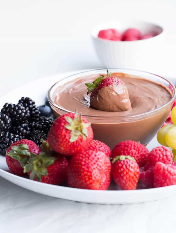 A bowl of fruit on a white plate with a strawberry being dipped in a chocolate spread