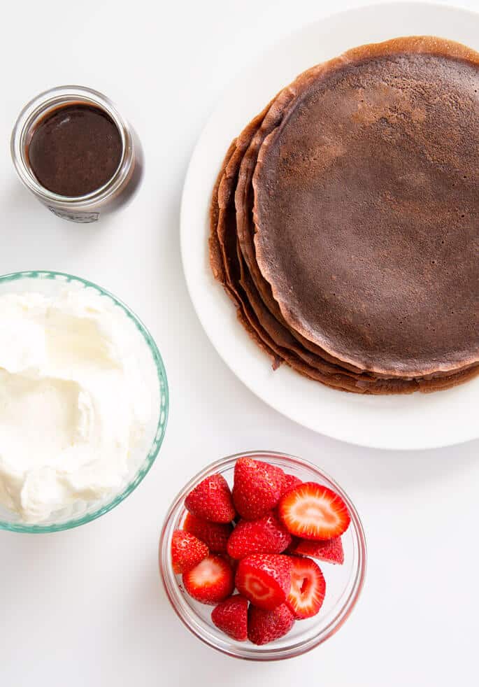 Overhead view of chocolate crepes and a bowl of whip cream, bowl of chocolate sauce and a bowl of strawberries 