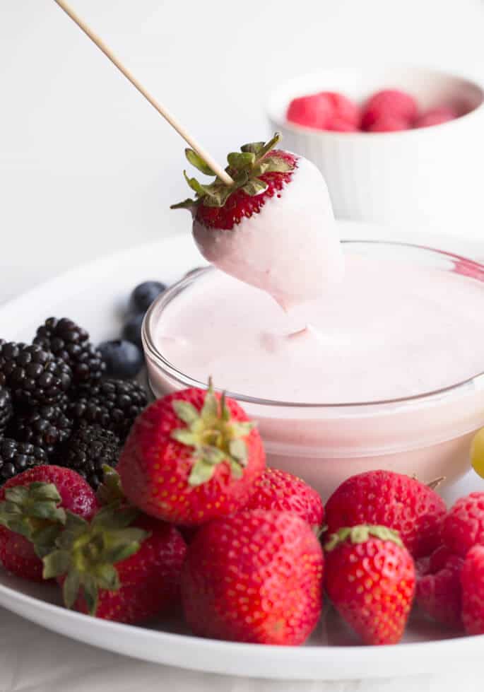 An assortment of fruit on a white plate with a strawberry on a wooden skewer being dipped in pink fruit dip in a small glass bowl