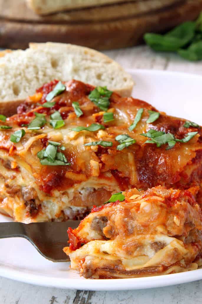 Lasagna with herbs on top on a white plate being scooped with a metal spatula 