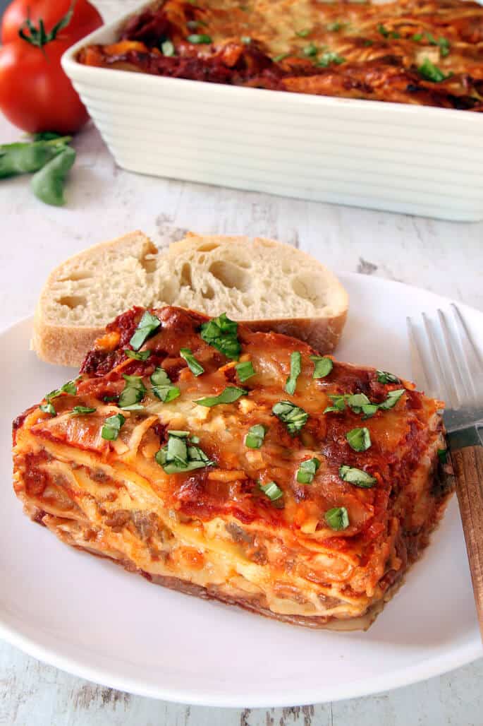 A close up of lasagna and a piece of bread on a white plate with a fork