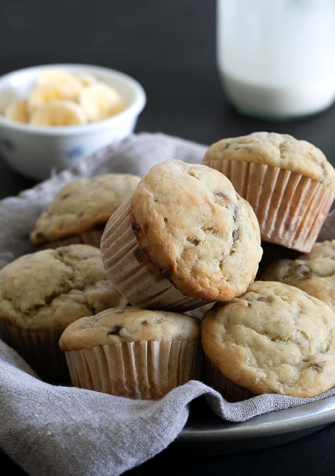 A pile of 7 or 8 light brown topped baked muffins with white muffin liners in a flat bowl lined with light tan cloth