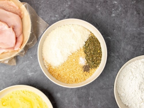 light brown breadcrumbs, white ground Parmesan cheese, white salt, black pepper, and green and brown dried Italian seasoning in wide flat light tan bowl