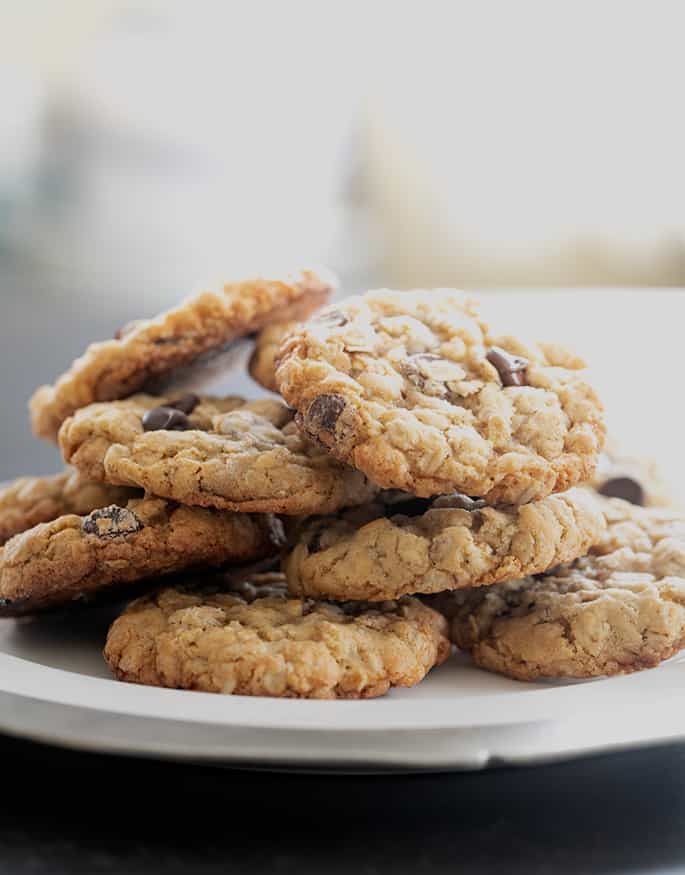 A plate of 8 baked cookies with oats and chocolate chips in a haphazard pile on a white serving platter with a metal edge