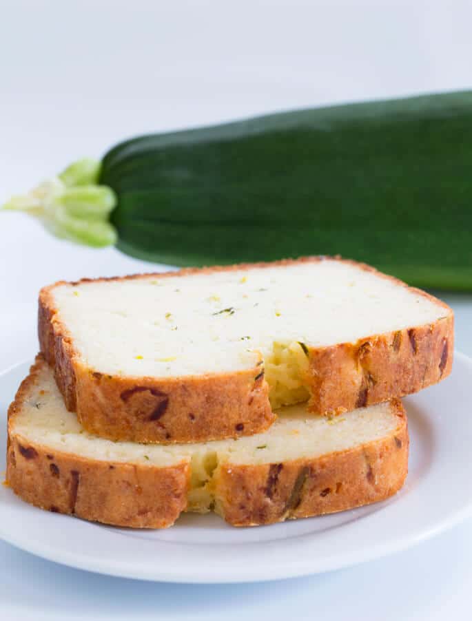 A stack of 2 slices of zucchini bread on a white plate and a zucchini in the back