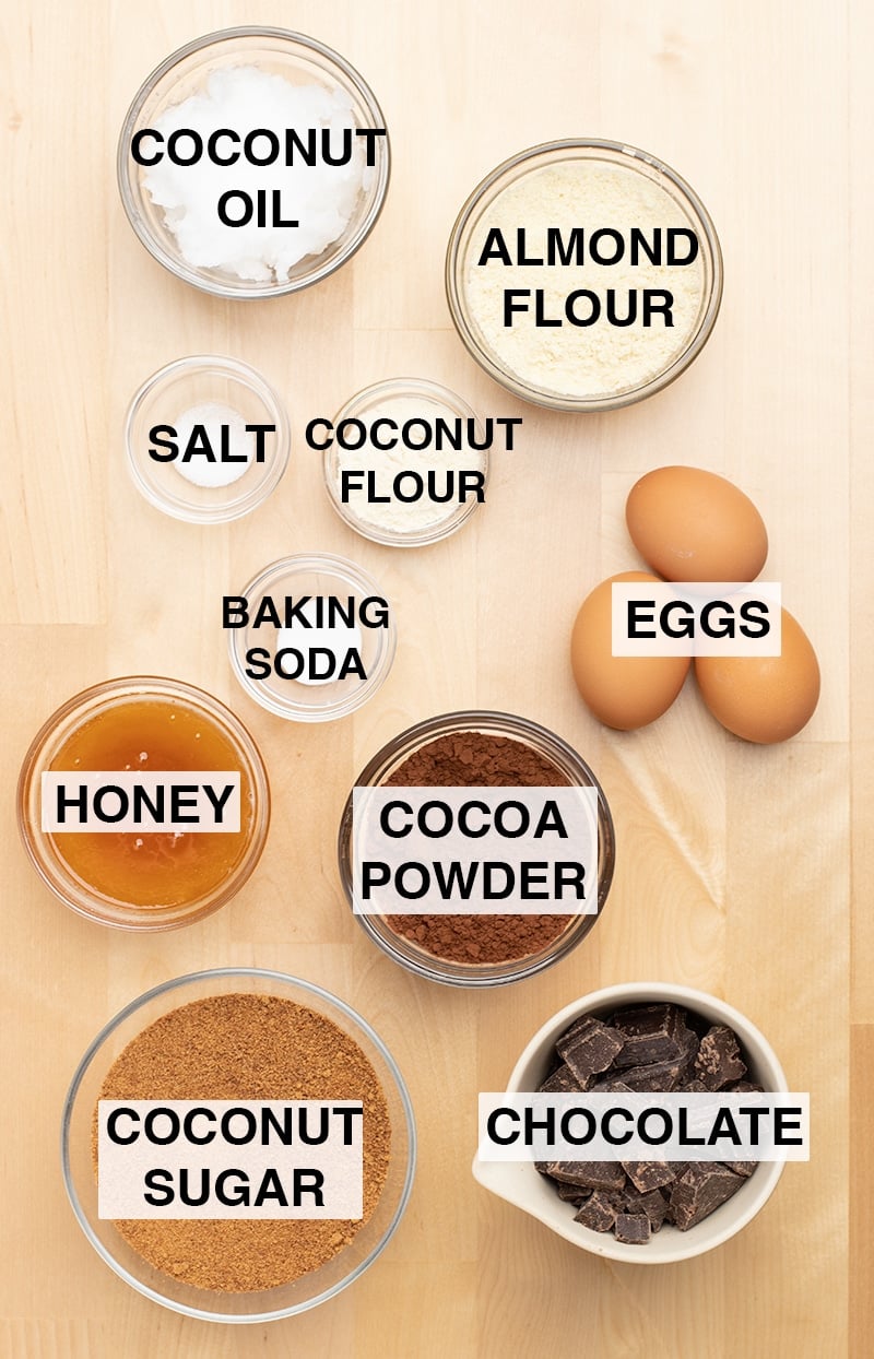 Almond flour, salt, coconut flour, honey, baking soda, eggs, cocoa powder, coconut sugar, chocolate, coconut oil in small glass bowls on blond wood table