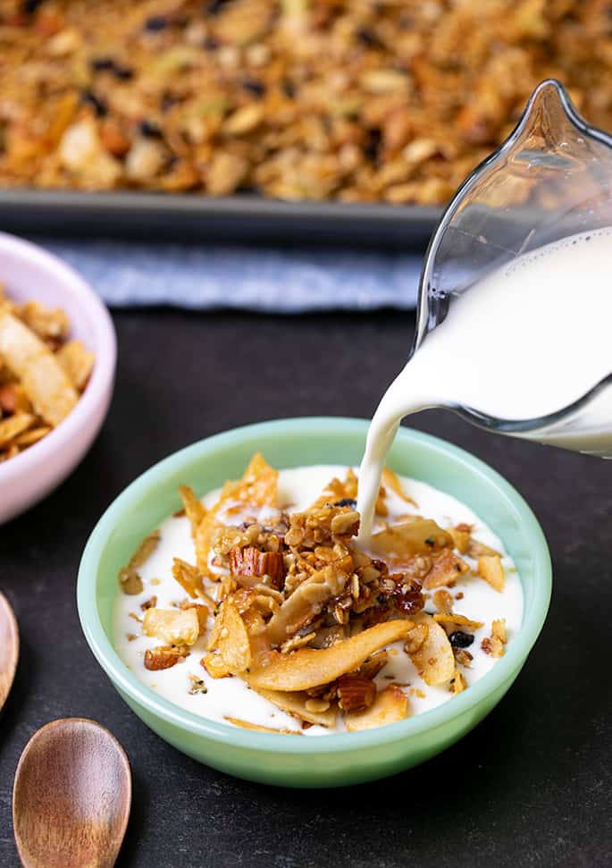 Milk pouring from glass jar into granola in small green bowl with spoons and pink bowl