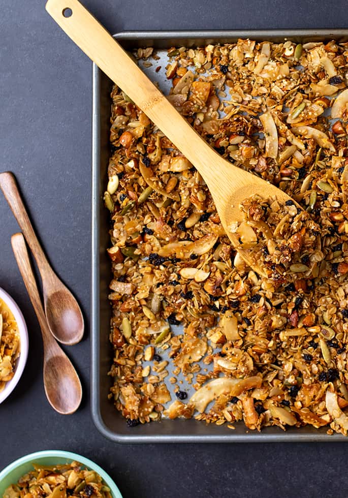 Overhead image of granola in rimmed baking sheet with wooden mixing spoon and individual wooden spoons on black surface