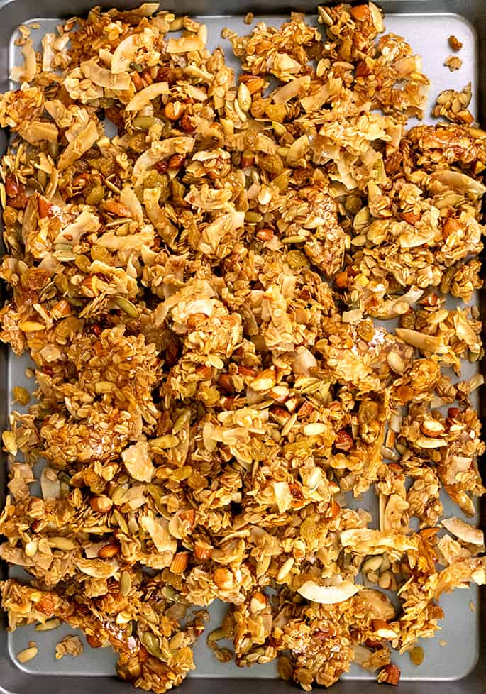 Overhead image of brown clusters of oats, nuts, seeds, and coconut chips on nonstick metal half sheet pan