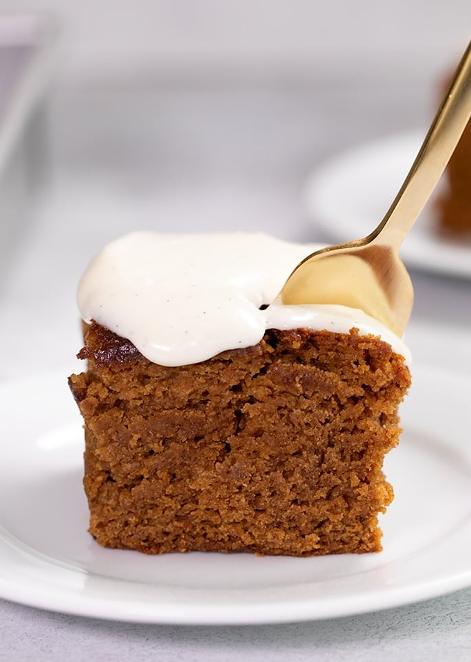 Fork placed in square gingerbred cake with frosting on a small white plate