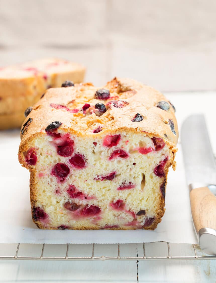 light yellow loaf of gluten free cranberry bread with light brown crust sliced in half on white paper lining wire rack with large bread knife