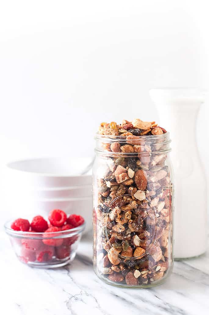 A jar of granola, and white bottle, white bowls and a bowl of raspberries on a white background

