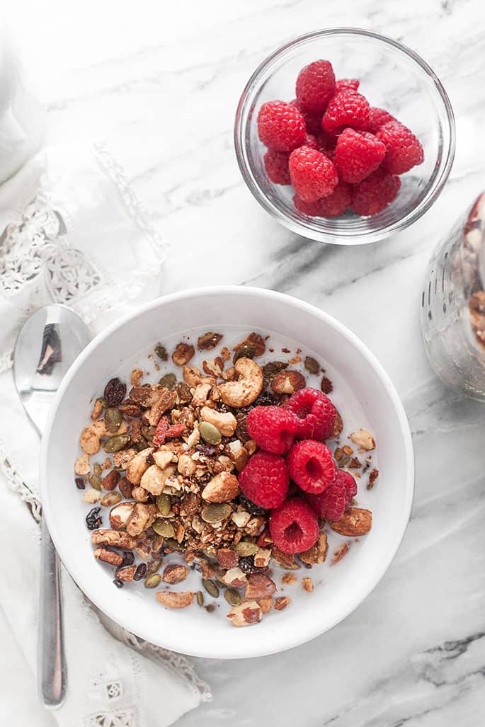 A bowl of granola with milk and raspberries with a spoon and a white cloth on a marble counter with a bowl of raspberries and jar of granola in background