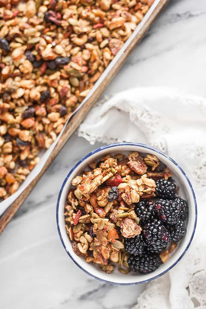 A bowl of granola with blackberries on a marble tabletop with white cloth on it and a tray of granola in corner of image