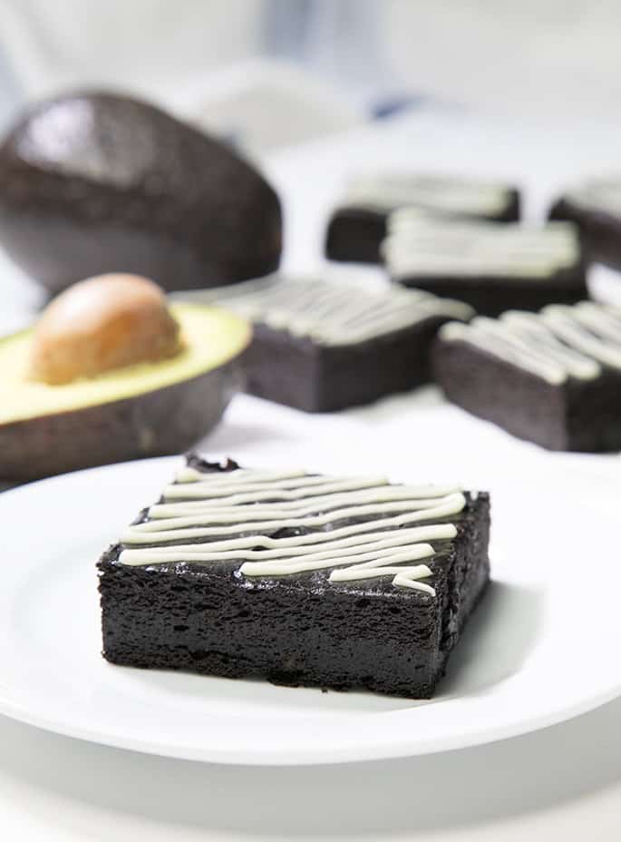 A avocado brownie with white zigzag pattern on top on a white plate with avocados and 5 avocado brownies with white zigzag pattern on top in white background