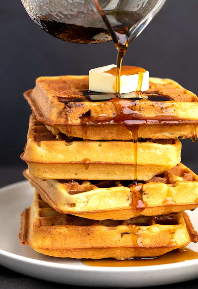 Syrup being poured on stack of 4 waffles with butter on top on small white plate