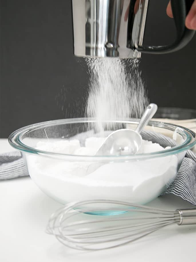 rice flour coming out of a handheld metal flour sifter into a glass bowl with rice flour and a metal dry ingredient scoop