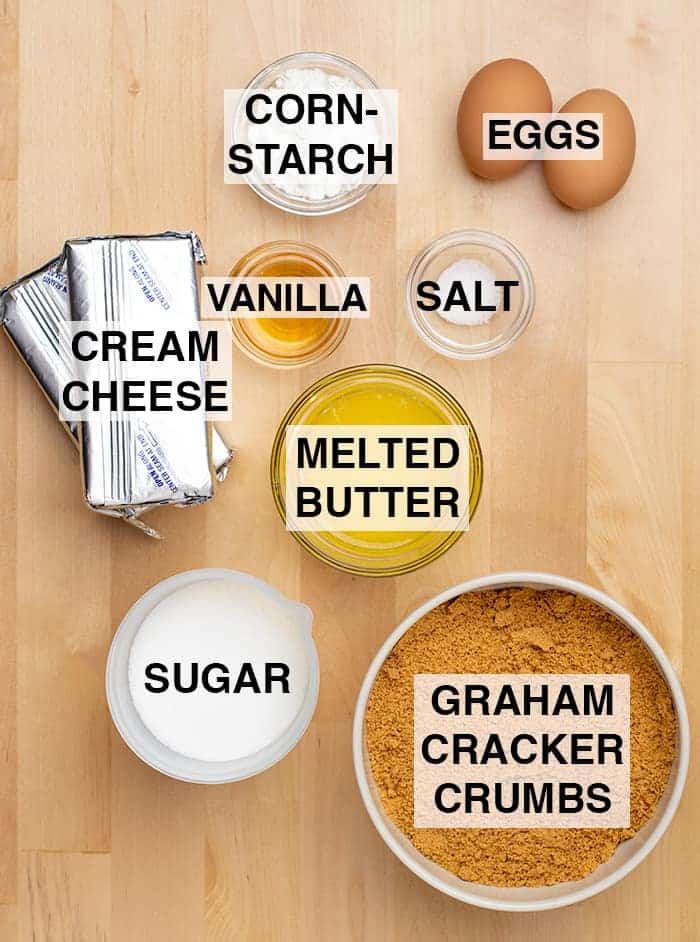 Ingredients in small bowls on light wood background, with identifying words: cornstarch, 2 eggs, 2 8 ounce foil packets of block cream cheese, vanilla, salt, white sugar, melted yellow butter, brown cookie crumbs