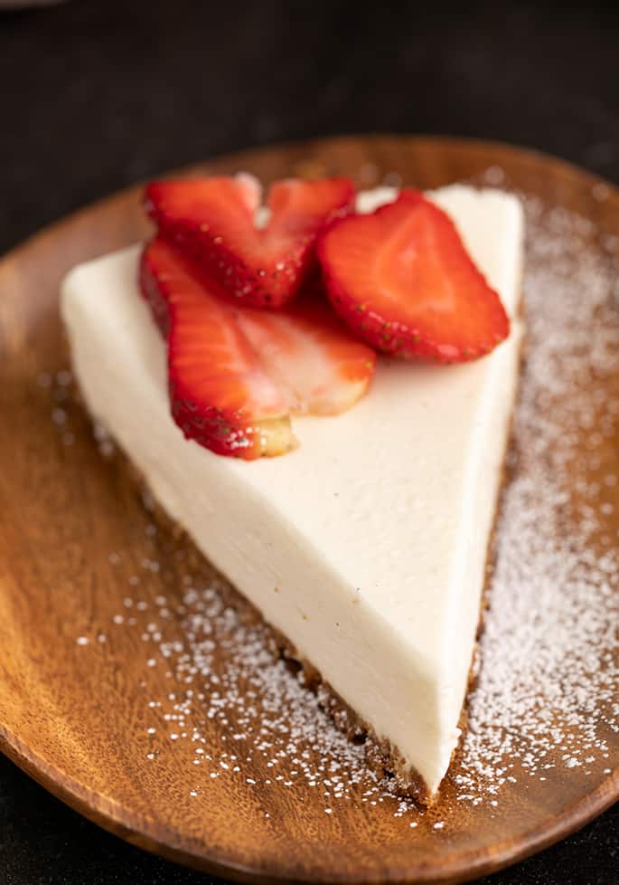 One slice of cheesecake with cookie crust with sliced strawberries and powdered sugar on a brown acacia wood plate