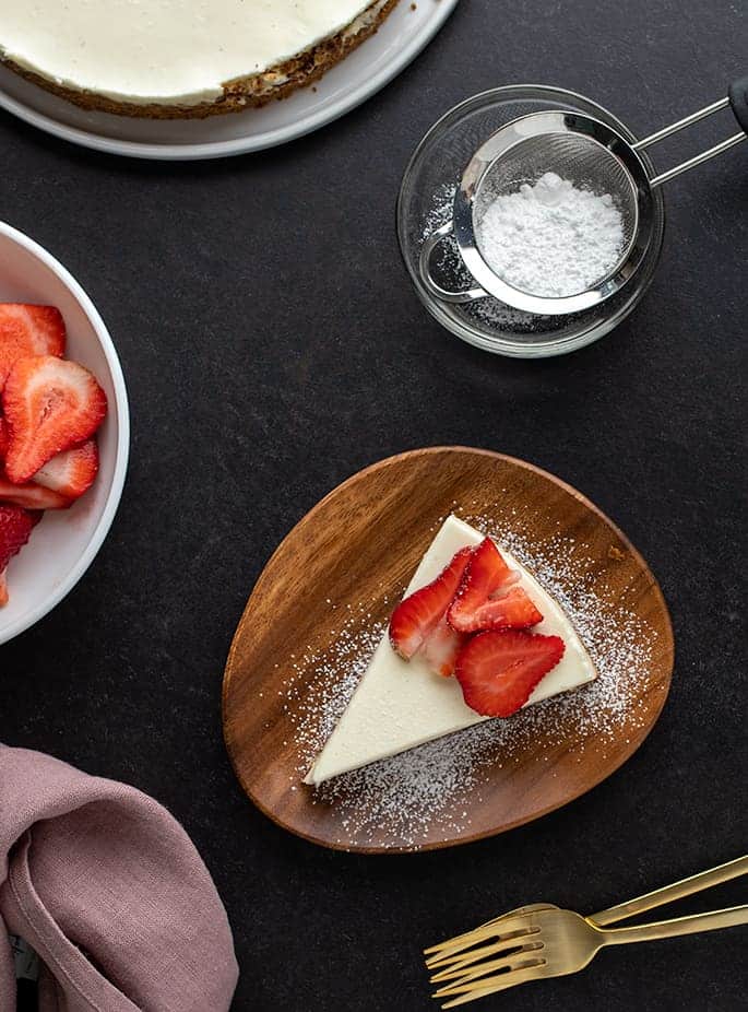 Overhead image of slice of cheesecake on small brown plate, sugar in sieve, rest of whole cheesecake, forks, and strawberries