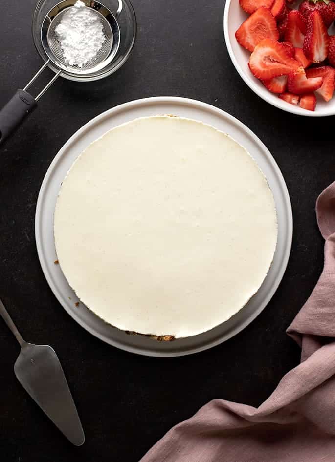 Overhead image of whole cheesecake on white platter with purple cloth, sliced strawberries in bowl, and powdered sugar in sieve