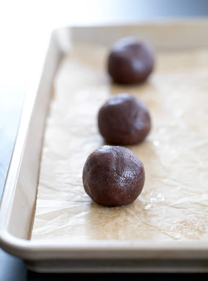 Raw balls of dark brown chocolate cookie dough on light brown paper on a metal baking sheet