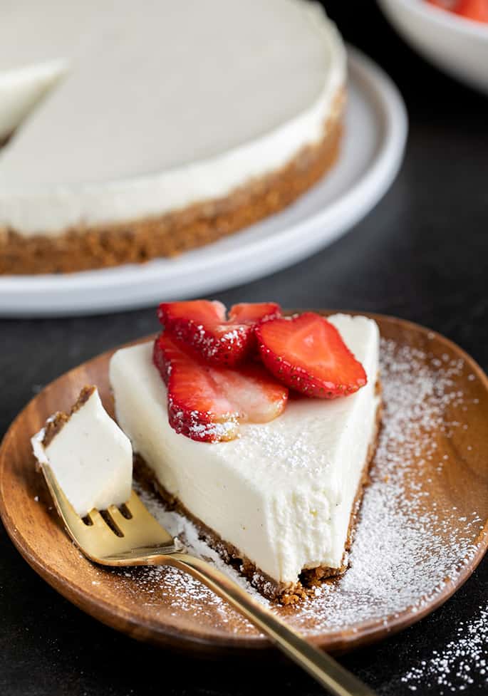 Slice of cheesecake with sliced strawberries and a forkful taken with the fork on a brown plate