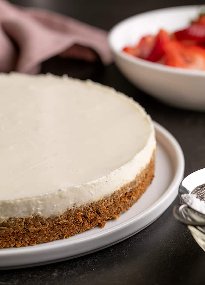 Whole cheesecake with cookie crust from side with sliced strawberries in white bowl in background