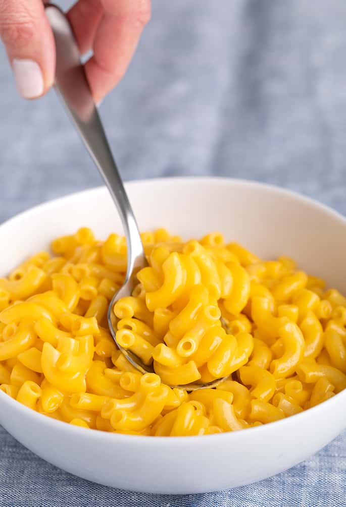 Fingers holding spoon taking a spoonful of elbow macaroni in cheese sauce in white bowl on blue cloth