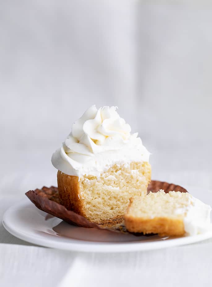 One gluten free vanilla cupcake with swirled white frosting cut in two pieces on brown paper liner on a small white plate above a white cloth