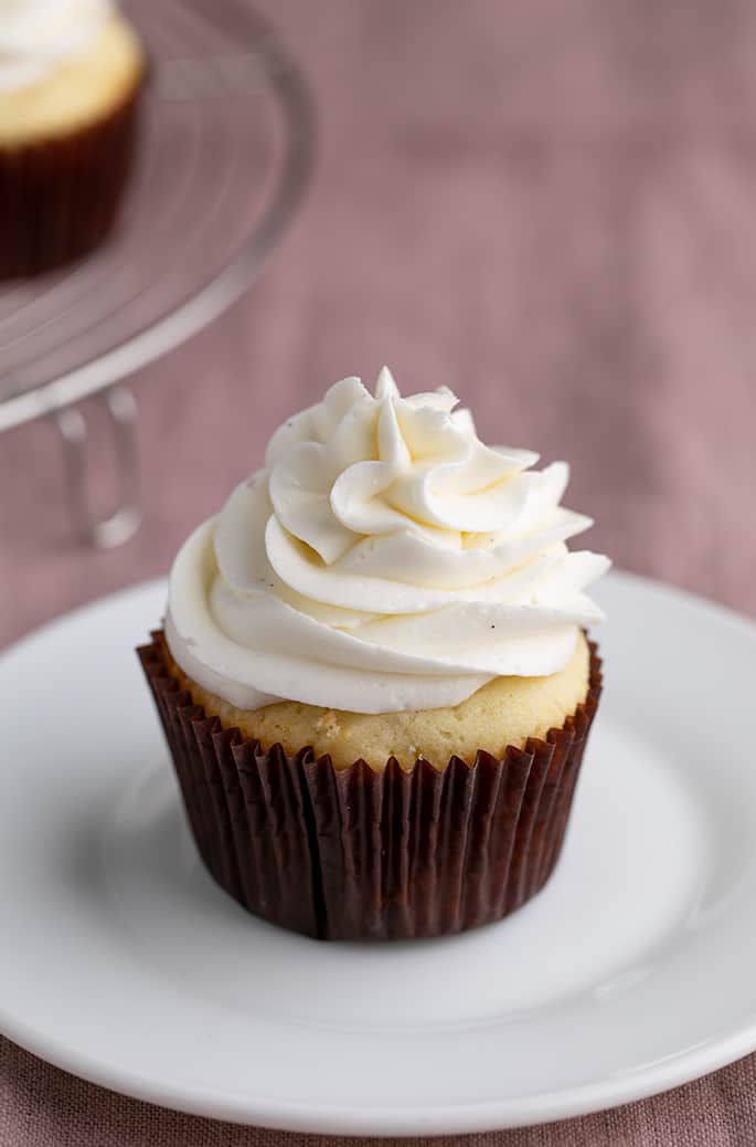 Vanilla cupcake in brown liner with white swirl of frosting on small white plate on purple cloth