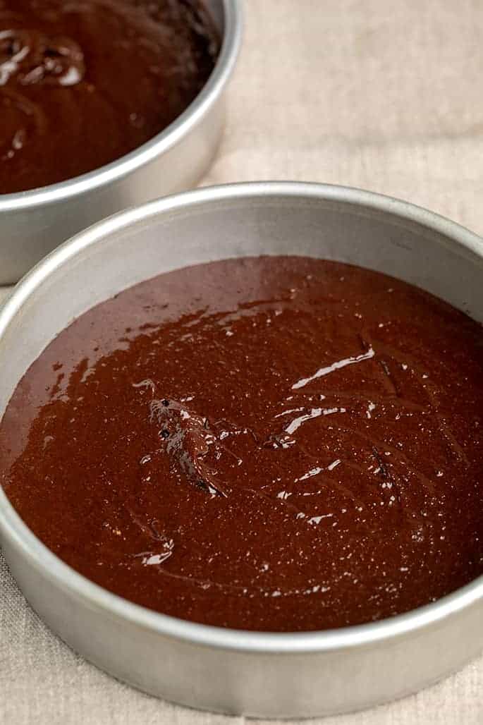 Raw chocolate cake batter in two round cake pans on tan cloth