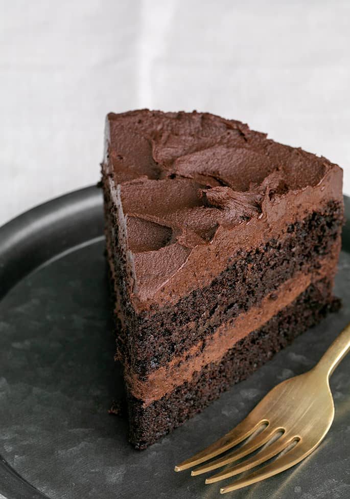 Slice of 2 layer frosted chocolate cake with fork on small dark plate on white cloth