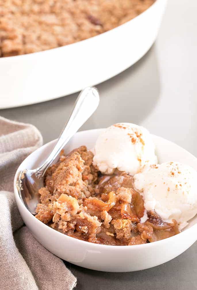 Brown cooked apples and crumb topping with two scoops of white ice cream with cinnamon sprinkled on top in a white bowl with a silver spoon