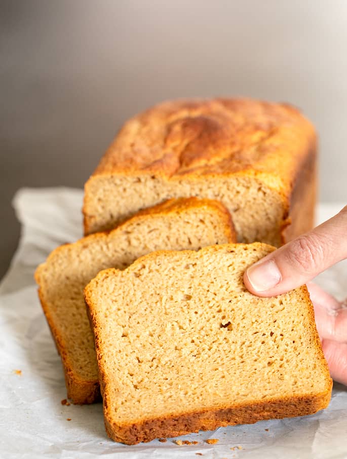 Hand taking a slice of light brown bread from a partially sliced loaf on white paper with crumbs