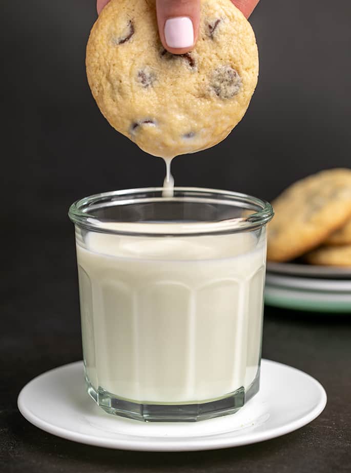 Fingers pulling out chocolate chip cookie from glass of milk on white plate with milk dripping off the cookie