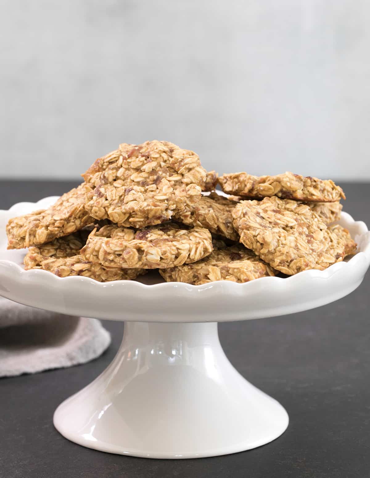 white footed cake plate with pile of light brown 3 ingredient oatmeal cookies on black surface with tan cloth
