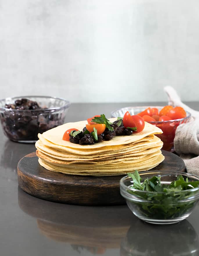 round brown wooden disk with stack of about 20 round light yellow tortillas topped with black beans, tomatoes and cilantro with more toppings in glass bowls