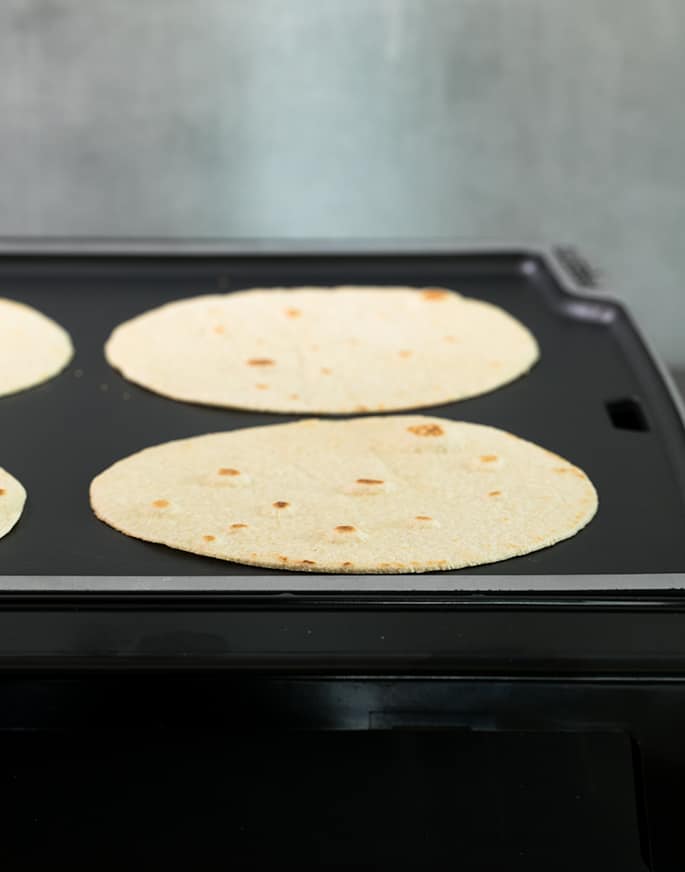 4 cooked light yellow tortilla rounds with browned spots on black electric skillet