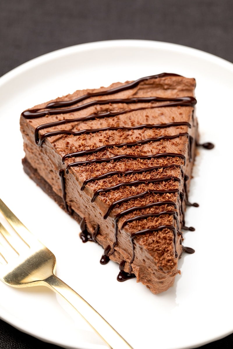 Closeup of slice of light brown cake with chocolate shavings and chocolate drizzle on white plate