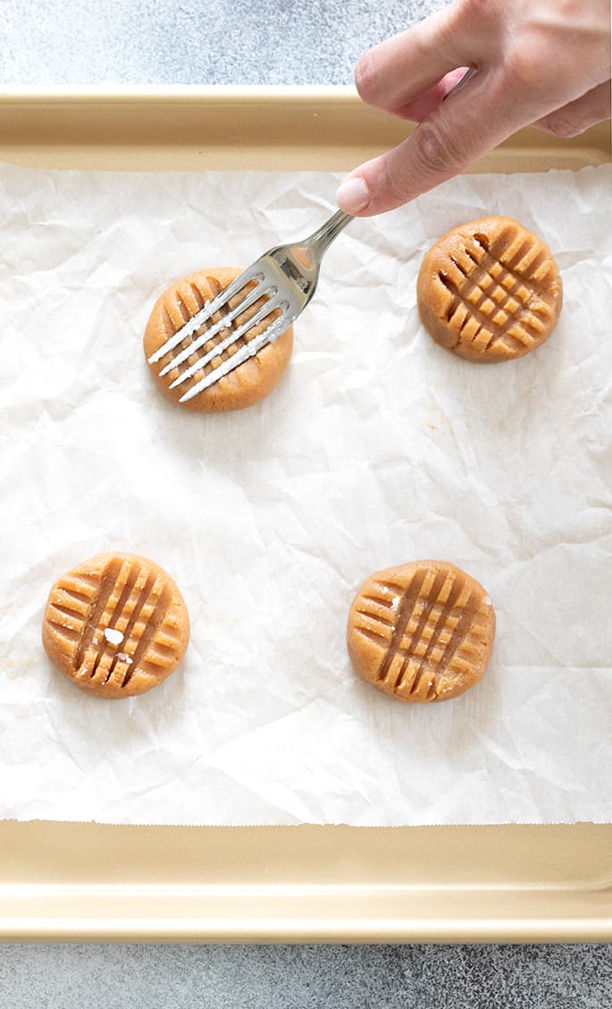 Hand pressing a fork into cookie dough on white paper on gold colored baking sheet