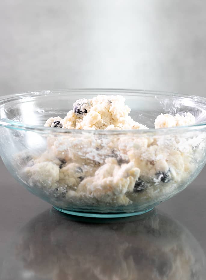 Large glass mixing bowl with pale raw clumps of pastry dough with blueberries mixed on on a gray table