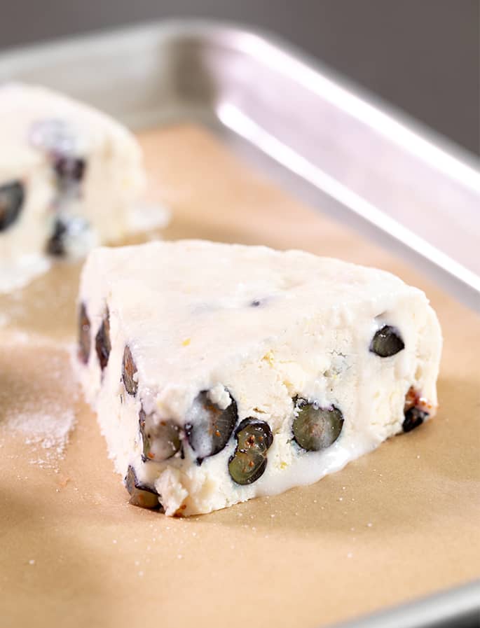 Closeup image of a pale raw triangle of pastry dough with blueberry halves in it on brown paper on a metal baking tray