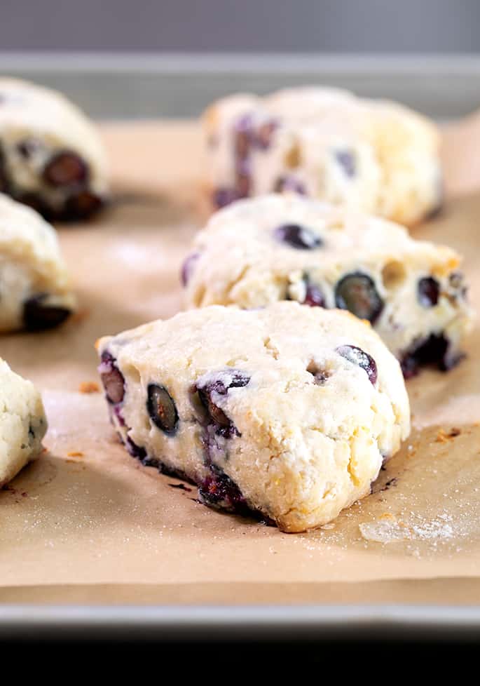 6 triangle shaped pale yellow pastries with blueberries on brown paper on a metal baking tray