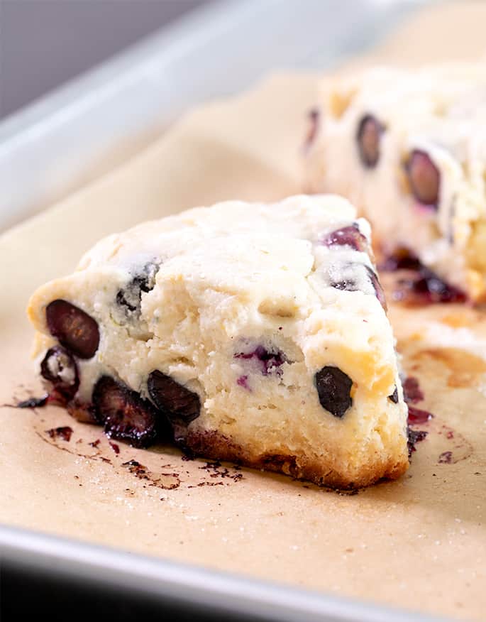 Closeup image of a light yellow baked triangle shaped pastry with blueberries on brown paper on a metal baking tray