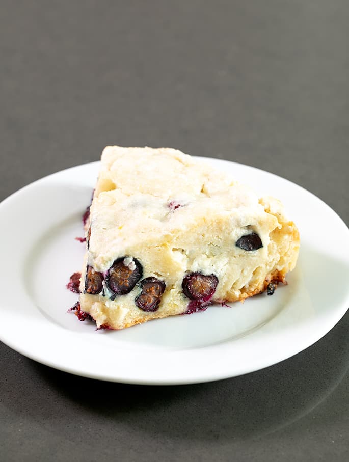 Pale yellow baked triangle with blueberry halves on small white plate on gray table