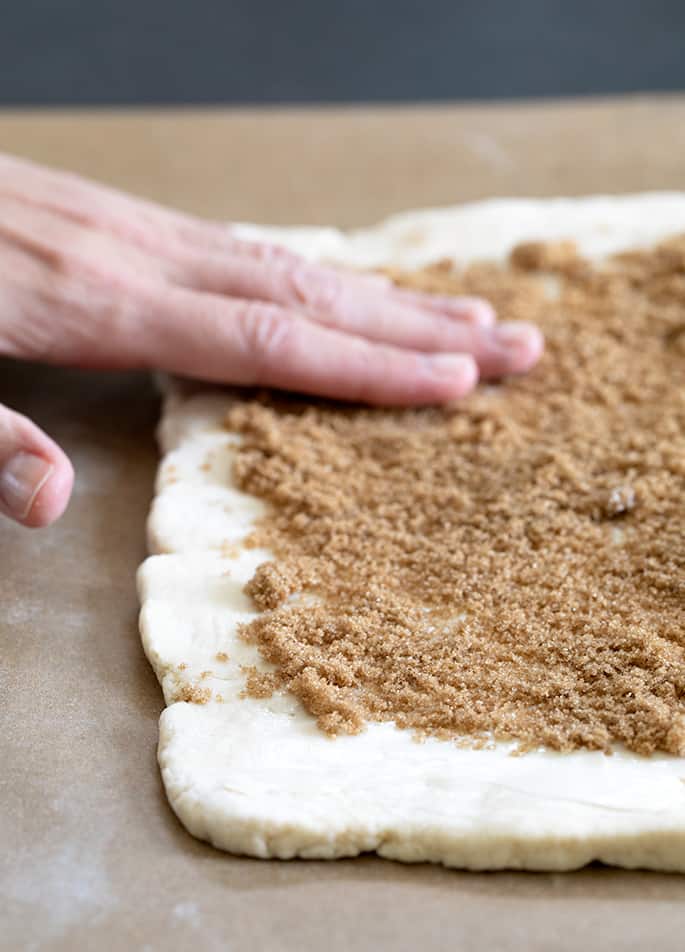 fingers of one hand pressing down on a thin layer of brown sugar on a rectangular shaped piece of white gluten free cinnamon roll dough on brown paper