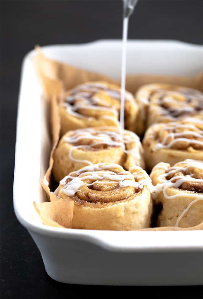 light brown coiled rolls with brown flecks packed tightly together in a white rectangular casserole dish lined with brown paper and white icing being drizzled on top