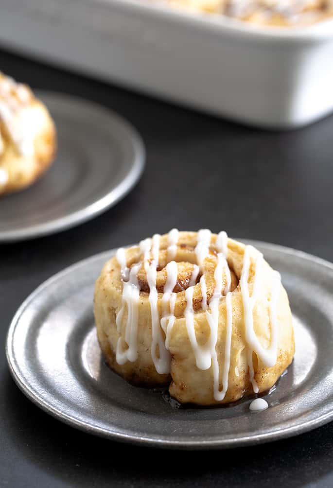 closeup image of a light brown coiled spiral roll with brown flecks and white drizzled icing on a small round metal plate