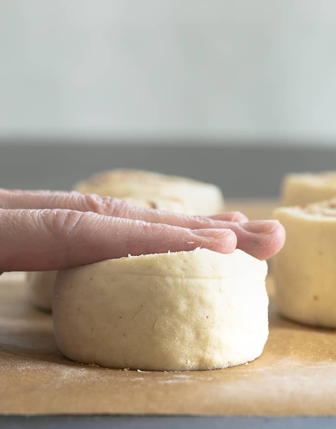 fingers pressing down on the top of a a round piece of white raw gluten free cinnamon roll dough on brown paper